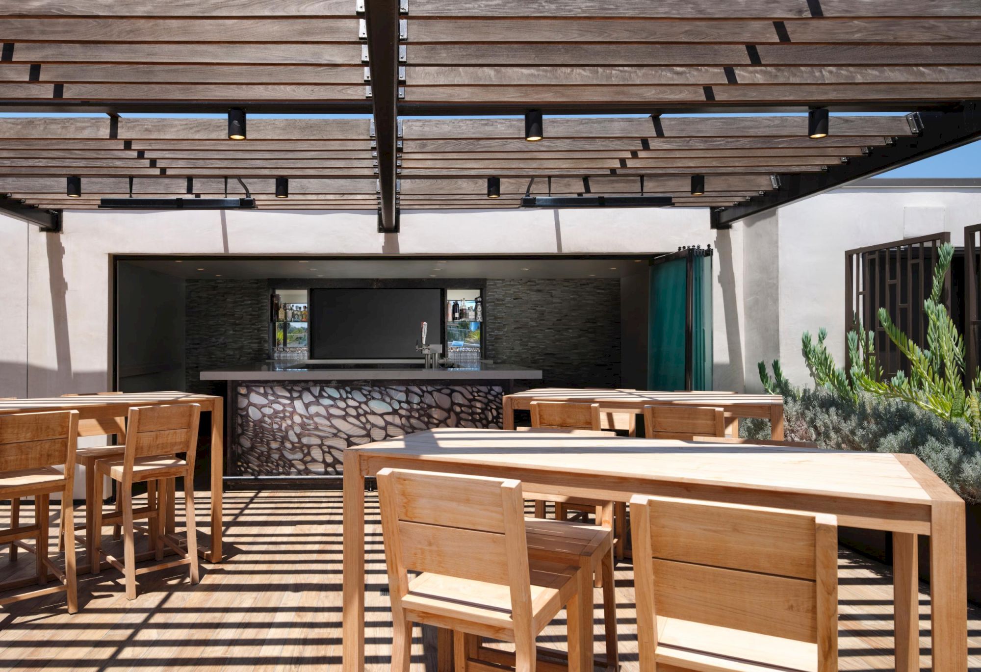 Outdoor seating area under a pergola with wooden tables and chairs, facing a bar with a modern design backdrop.