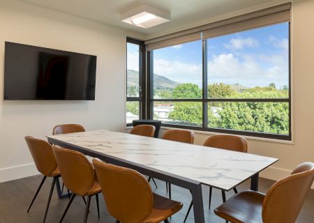 A modern conference room with a marble table, brown chairs, a wall-mounted TV, and large windows with a scenic view outside.