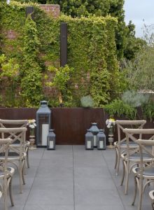 Outdoor seating area with wooden chairs arranged in rows, a backdrop of greenery, and lanterns on the ground.