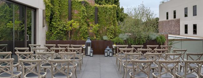 Outdoor seating area with wooden chairs arranged in rows, a backdrop of greenery, and lanterns on the ground.