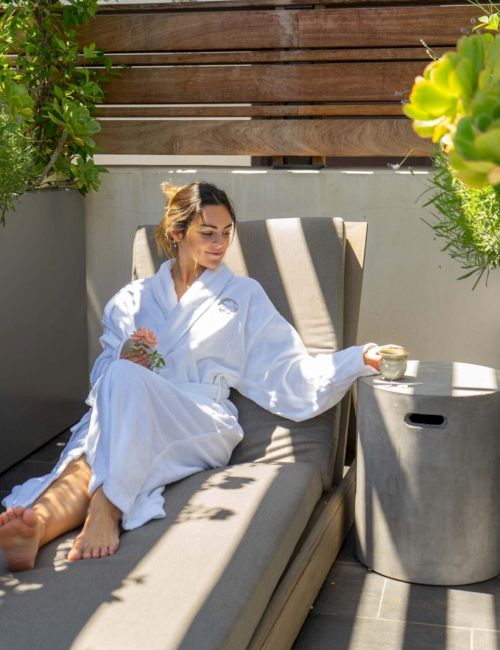 A person relaxes in a robe on a lounge chair outdoors, holding a flower and a cup, surrounded by plants and sunlight.