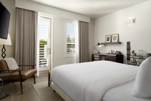 A modern hotel room with a bed, TV, chair, desk with a lamp, and a minibar. Bright lighting and window with curtains and blinds.