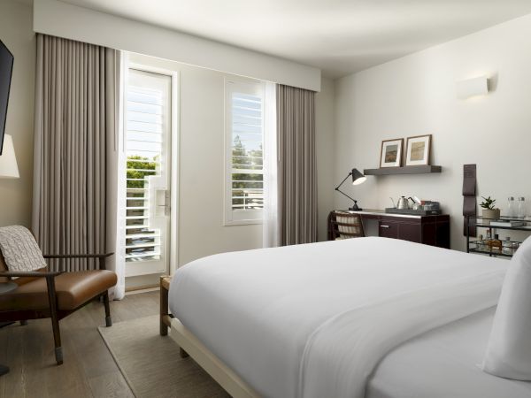 A modern hotel room with a bed, TV, chair, desk with a lamp, and a minibar. Bright lighting and window with curtains and blinds.