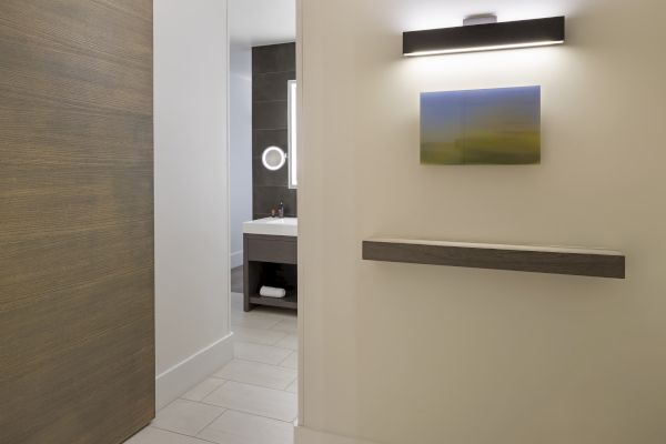 A modern bathroom with dark tiles, a mirror with a light, a sink, and a small towel shelf. There's also abstract wall art and a light above.