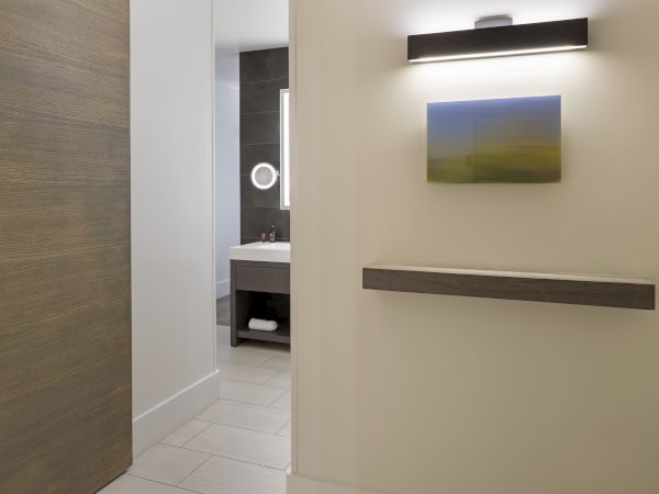 A modern bathroom with dark tiles, a mirror with a light, a sink, and a small towel shelf. There's also abstract wall art and a light above.