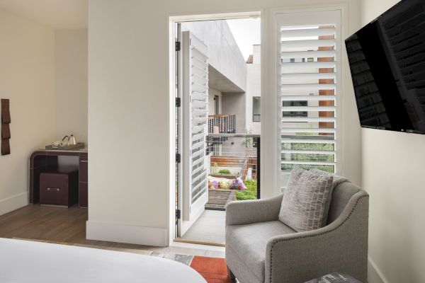 A modern room with a gray chair, wall-mounted TV, and an open door leading to a balcony with garden views outside.