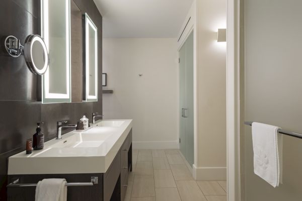A modern bathroom features a double sink vanity, backlit mirrors, and a towel hanging by the shower area.