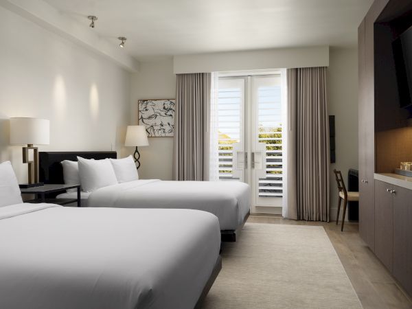 A modern hotel room with two beds, a lamp, artwork, and a window with curtains. A TV and furniture are also visible in the room.