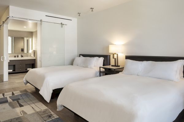 The image shows a hotel room with two white beds, a lamp, and a visible bathroom with a sliding glass door.