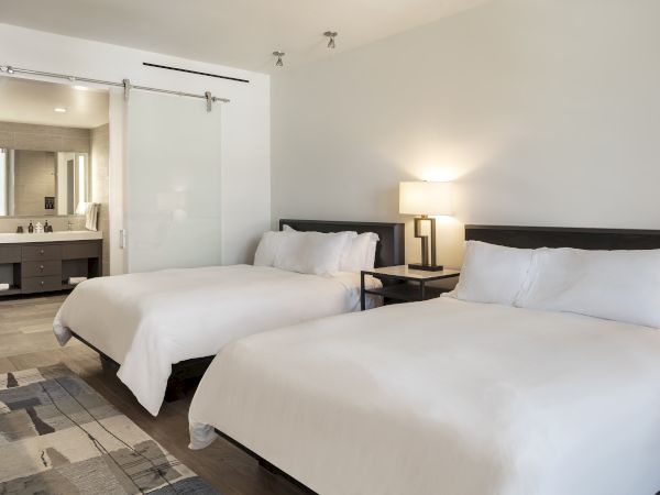 The image shows a hotel room with two white beds, a lamp, and a visible bathroom with a sliding glass door.