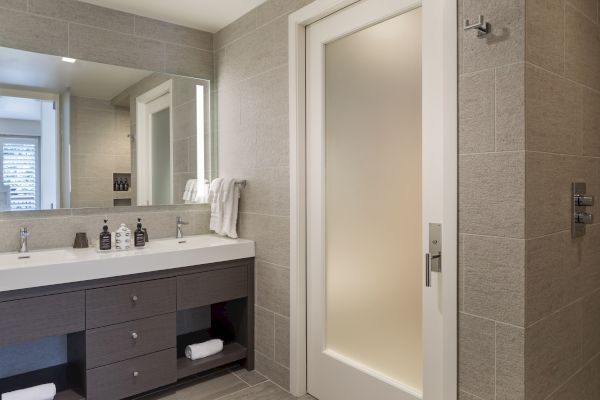A modern bathroom with a large mirror, double sink, towels, and toiletries on the counter. There's a frosted glass door visible.