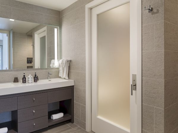 A modern bathroom with a large mirror, double sink, towels, and toiletries on the counter. There's a frosted glass door visible.