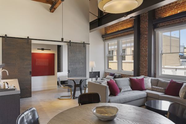 The image shows a modern living area with a large sofa, dining table, barn doors, and windows letting in natural light, featuring stylish decor.