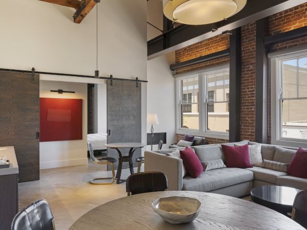 The image shows a modern living area with a large sofa, dining table, barn doors, and windows letting in natural light, featuring stylish decor.