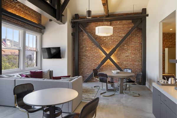 A modern loft-style room with brick walls, a sectional sofa, round tables, chairs, a mounted TV, and ceiling beams.