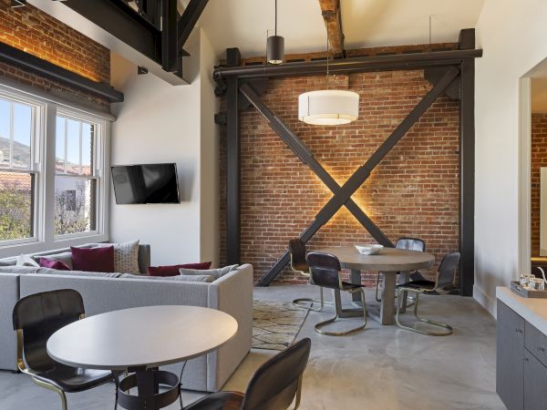 A modern loft-style room with brick walls, a sectional sofa, round tables, chairs, a mounted TV, and ceiling beams.