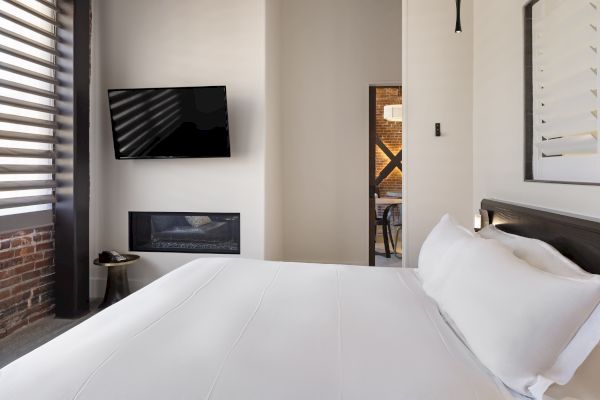 The image shows a modern bedroom with a bed, wall-mounted TV, fireplace, and a glimpse into another room.