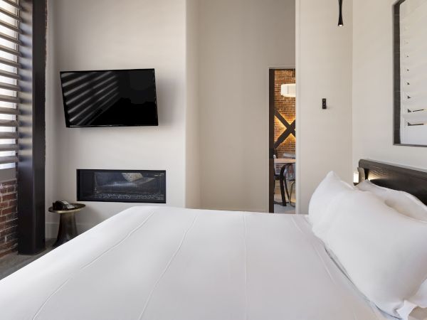 The image shows a modern bedroom with a bed, wall-mounted TV, fireplace, and a glimpse into another room.