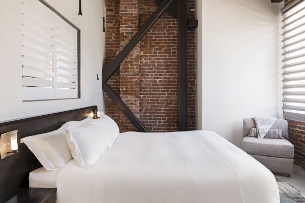 A cozy bedroom with a white bed, brick wall, and an armchair with a pillow and blanket. A shuttered window and modern lights are also visible.