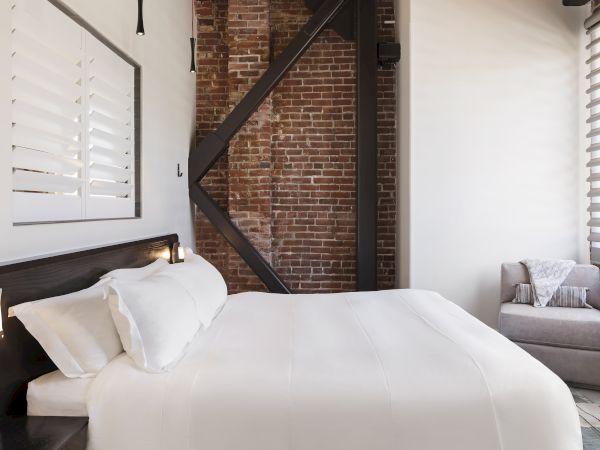 A cozy bedroom with a white bed, brick wall, and an armchair with a pillow and blanket. A shuttered window and modern lights are also visible.