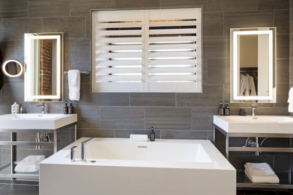 A modern bathroom with a white tub, twin sinks with mirrors, towels, and gray tile walls. Simple and elegant design.