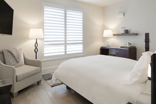 A cozy bedroom with a bed, armchair, floor lamp, window shutters, TV, and a shelf with a kettle and glasses.
