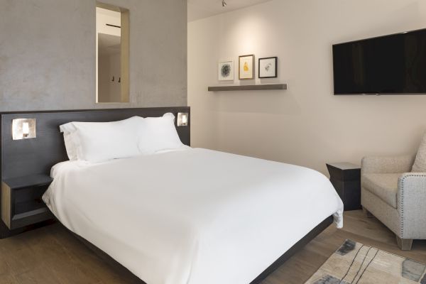A modern bedroom with a double bed, wall art, a TV, and an armchair. The room has a minimalist and elegant decor style.