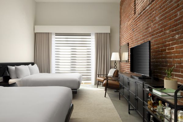 A hotel room with two beds, brick wall, TV, chair, lamp, and a minibar cart near a window with curtains.