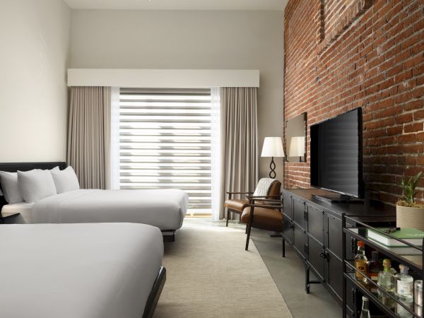 A hotel room with two beds, brick wall, TV, chair, lamp, and a minibar cart near a window with curtains.