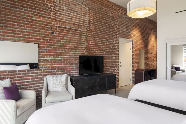 A modern room with exposed brick walls, two beds, chairs, a TV, and soft lighting.