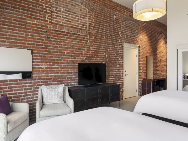 A modern room with exposed brick walls, two beds, chairs, a TV, and soft lighting.