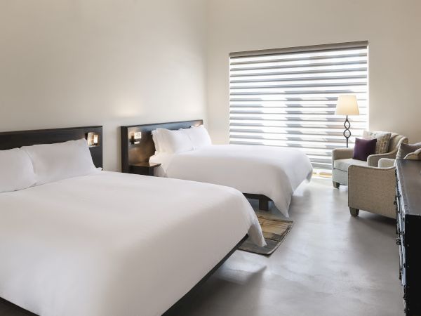 A hotel room with two large beds, an armchair, side table, lamp, TV, and window blinds allowing natural light to enter.