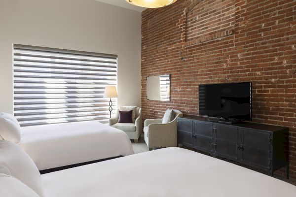 A hotel room with exposed brick, two beds, a TV on a black stand, and a cozy seating area with a lamp.