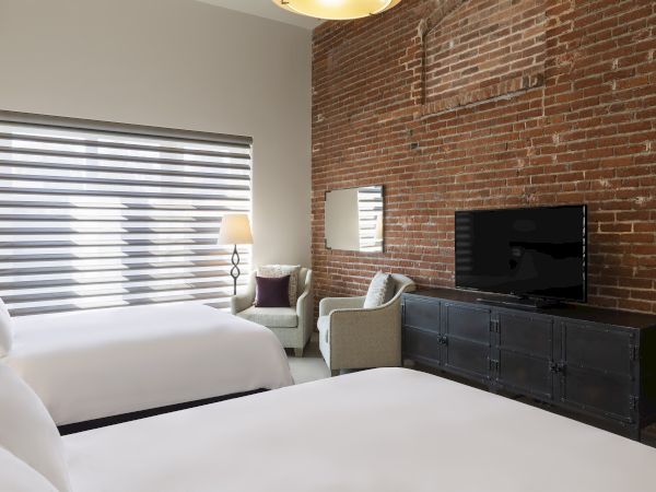 A hotel room with exposed brick, two beds, a TV on a black stand, and a cozy seating area with a lamp.