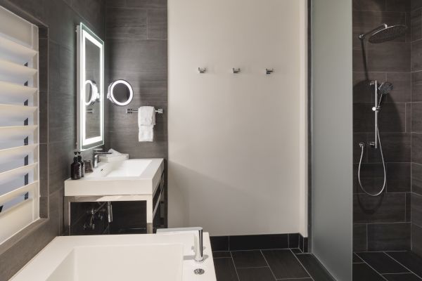 A modern bathroom with a lit mirror, sink, towel rack, and a shower featuring a rainfall showerhead with dark tile walls.