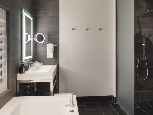 A modern bathroom with a lit mirror, sink, towel rack, and a shower featuring a rainfall showerhead with dark tile walls.
