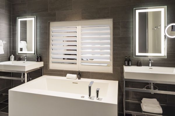 Luxurious bathroom with a large tub, two sinks, backlit mirrors, and window shutters, featuring modern design elements throughout.