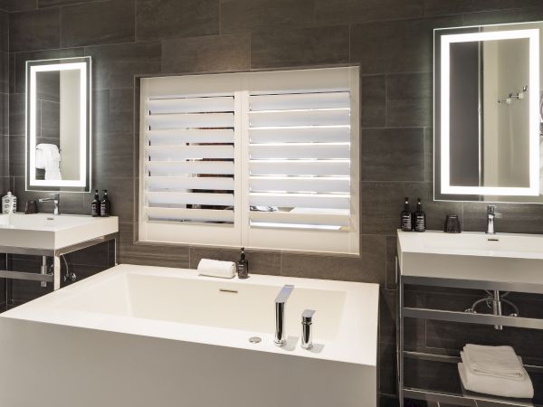 Luxurious bathroom with a large tub, two sinks, backlit mirrors, and window shutters, featuring modern design elements throughout.