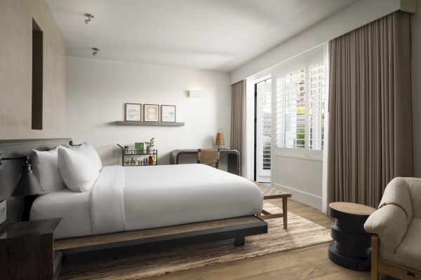 A modern bedroom with a large bed, small desk, artwork, and bright window with shutters and curtains.