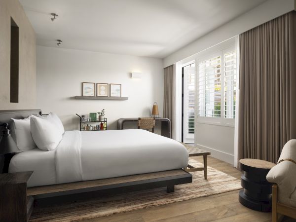 A modern bedroom with a large bed, small desk, artwork, and bright window with shutters and curtains.