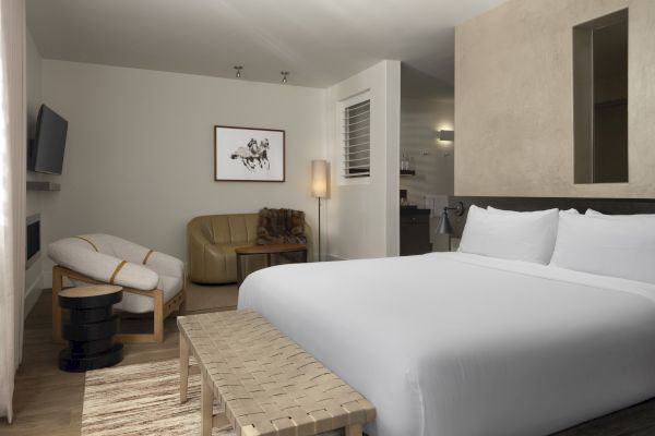 A modern, stylish hotel room with a bed, seating area, artwork, and soft lighting, featuring neutral tones and sleek furnishings.