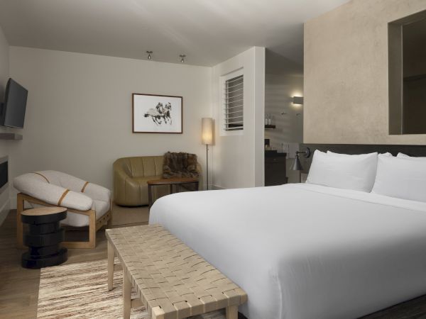 A modern, stylish hotel room with a bed, seating area, artwork, and soft lighting, featuring neutral tones and sleek furnishings.