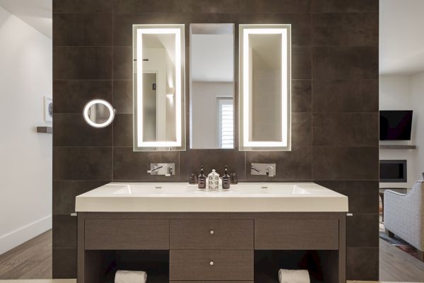 Modern bathroom with a double sink, two backlit mirrors, and a dark tile wall. It includes a magnifying mirror and toiletries.