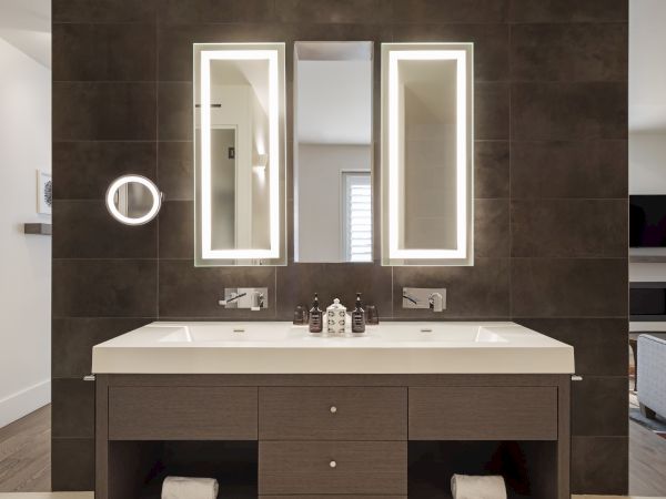Modern bathroom with a double sink, two backlit mirrors, and a dark tile wall. It includes a magnifying mirror and toiletries.