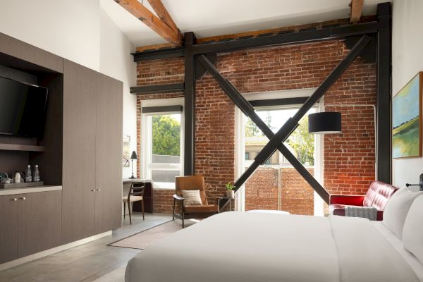 A modern bedroom with exposed brick walls, wooden beams, a large bed, desk, leather chair, and a wall-mounted TV.