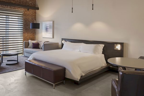 A modern bedroom with a large bed, sofa, round table, and chairs. Minimalist design with neutral tones and exposed brick wall.