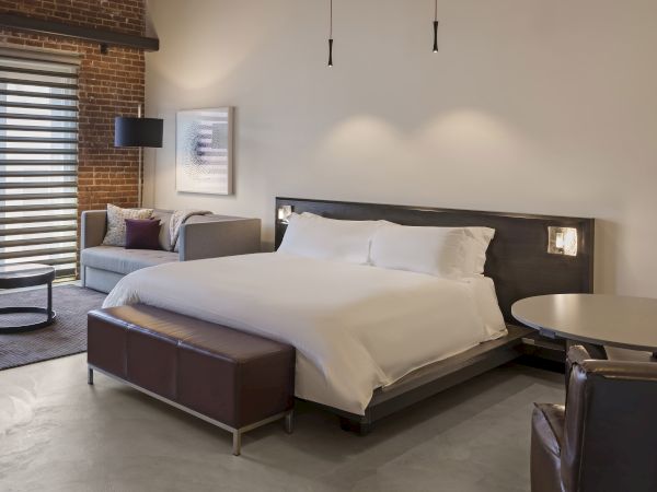 A modern bedroom with a large bed, sofa, round table, and chairs. Minimalist design with neutral tones and exposed brick wall.