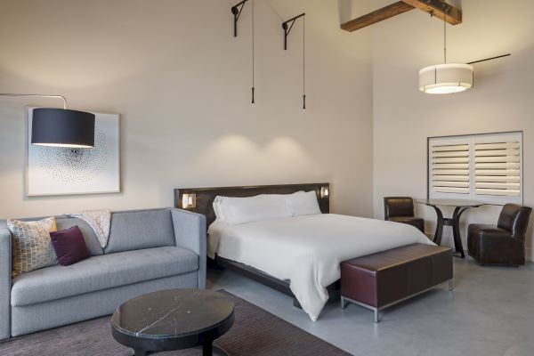 A modern bedroom with a bed, sofa, table, chairs, and artwork on the wall. Neutral colors and soft lighting enhance the cozy ambiance.