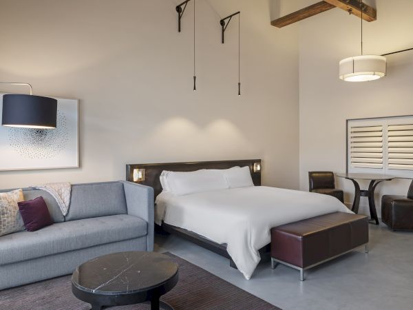 A modern bedroom with a bed, sofa, table, chairs, and artwork on the wall. Neutral colors and soft lighting enhance the cozy ambiance.