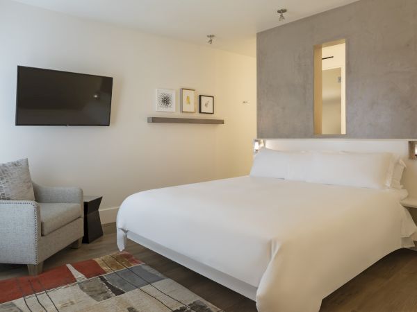 A modern bedroom with a bed, gray chair, wall-mounted TV, and abstract art on the wall.